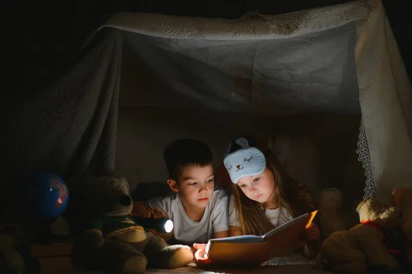 子供のテントで読書や家族のゲームをしています ベッドに行く前に本や懐中電灯を持っている男の子と女の子 — ストック写真