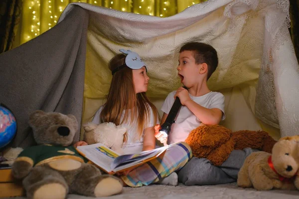 两个拿着手电筒的孩子在毛毯下看书当帐篷 — 图库照片