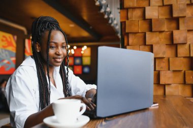 Genç Afrikalı Amerikalı kız restoranda oturuyor ve dizüstü bilgisayarıyla yazı yazıyor. Kafede bilgisayarla çalışan güzel bir kız..