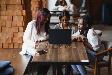 Üç Afrikalı Amerikalı kız (öğrenci) kafede oturmuş test için hazırlanıyor ya da ödevlerini birlikte yapıyor, dizüstü bilgisayar ve dijital tablet kullanıyorlar..