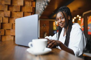 Kafede oturan, kahve içen ve dizüstü bilgisayarla çalışan genç bir kadın.