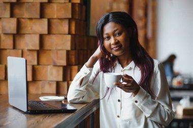 Kafede oturan, kahve içen ve dizüstü bilgisayarla çalışan genç bir kadın.