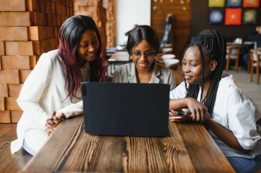 Üç Afrikalı Amerikalı kız (öğrenci) kafede oturmuş test için hazırlanıyor ya da ödevlerini birlikte yapıyor, dizüstü bilgisayar ve dijital tablet kullanıyorlar..