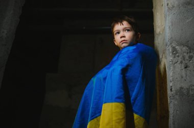 A sad Ukrainian boy wrapped in a Ukrainian flag, hiding from bombings in a bomb shelter in the city of Mariupol or Kiev. Russia's aggression. The war in Ukraine and 2022. clipart