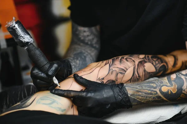 Cropped close up of a bearded tattoo artist working at his studio tattooing sleeve on the arm of his male client. Man getting tattooed by professional tattooist