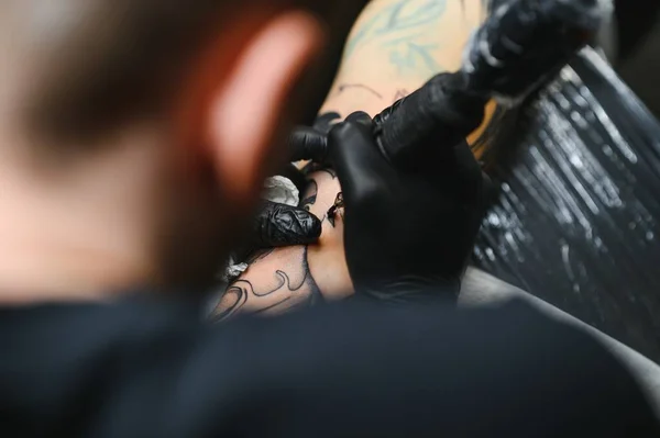 Cropped close up of a bearded tattoo artist working at his studio tattooing sleeve on the arm of his male client. Man getting tattooed by professional tattooist