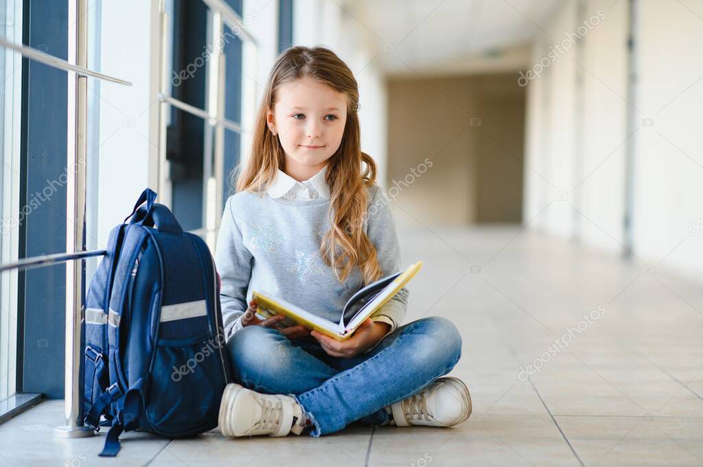 Little pretty girl sitting on the floor of the school hallway and reading a book. The concept of schooling.