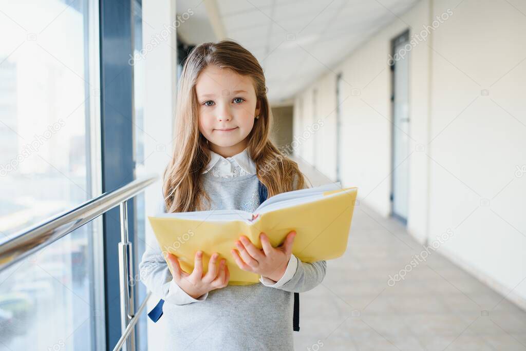 Front view of little beautiful school girl among corridor at school, holding notes at hands. Funny and happy girl smiling at camera, resting after lessons on primary school.