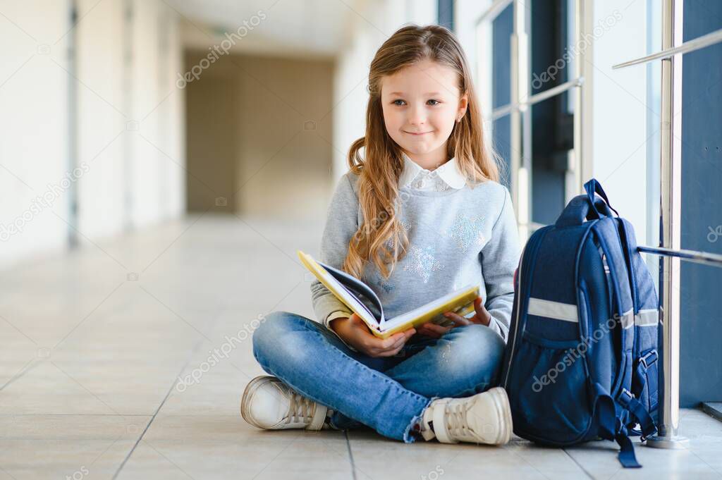 Little pretty girl sitting on the floor of the school hallway and reading a book. The concept of schooling.