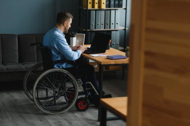 Özel ihtiyaçları olan, günlük giysiler içinde, kablosuz bilgisayarla çalışan genç bir adam. Tekerlekli sandalyede otururken evden çalışan serbest çalışan bir erkek..