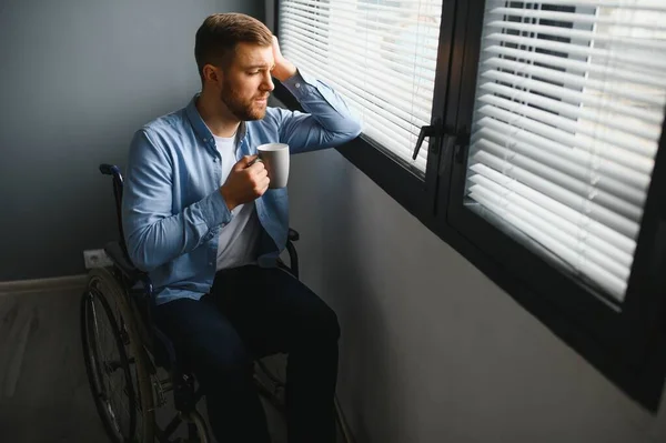 Sad disabled person feeling lonely at home or clinic. Depression on self isolation. Upset man in wheelchair looks out of large window and dreams, in living room interior