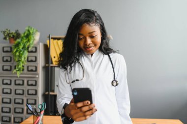 Gülümseyen Afrikalı doktor kadın doktor cep telefonuyla iş yerinde konuşuyor. Sağlık bakımı uzmanı telefon görüşmelerine cevap veriyor. Akıllı telefonlar üzerinde uzaktan danışmanlık yapıyor. Randevu notları alıyor.