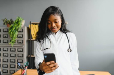 Gülümseyen Afrikalı doktor kadın doktor cep telefonuyla iş yerinde konuşuyor. Sağlık bakımı uzmanı telefon görüşmelerine cevap veriyor. Akıllı telefonlar üzerinde uzaktan danışmanlık yapıyor. Randevu notları alıyor.