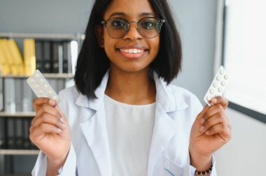 Gülümseyen Afrikalı kadın doktor ya da eczacının, renkli kabarcıkları haplarla tutarken, eczanede ya da hastanede kameraya gösterirken, yatay çekimini kapat.