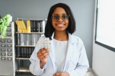 Gülümseyen Afrikalı kadın doktor ya da eczacının, renkli kabarcıkları haplarla tutarken, eczanede ya da hastanede kameraya gösterirken, yatay çekimini kapat.