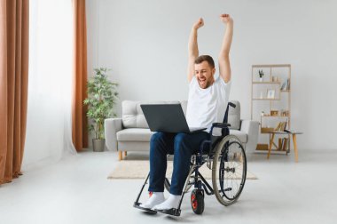 Gülümseyen engelli erkek portresi tekerlekli sandalyede oturuyor ve bilgisayarla evden çalışıyor. Özel ihtiyaçları olan genç bir işçi. Serbest Çalışanlar ve Engelliler Konsepti.