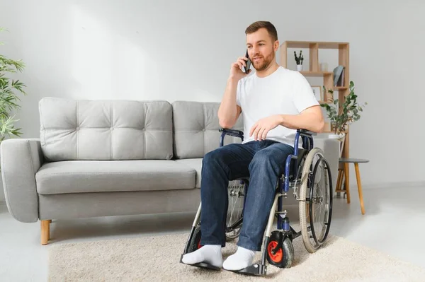 残疾人坐在轮椅上 他在用智能手机和别人说话 他在他那明亮的大客厅里 他笑了 — 图库照片