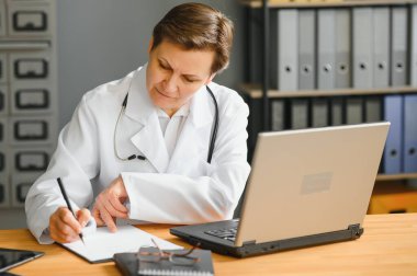 Orta yaşlı kadın doktorun portre fotoğrafı. Masada oturuyor ve ofisinde çalışıyor..