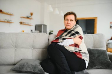 Orta yaşlı, 50 'li yaşlarda, hasta, kanepede oturan, sıcak ekoseli hapşıran, akıcı burunlu, mevsimsel soğuk, zayıf bağışıklık sistemi konsepti olan bir kadın.