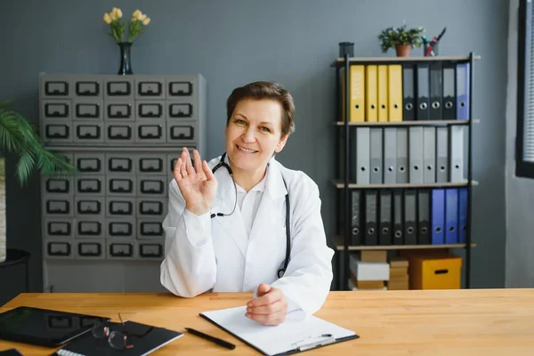 Portrait of female doctor counseling patient via video call. Professional physician in white lab coat gesturing and explaining course of treatment sitting at office desk during online consultation