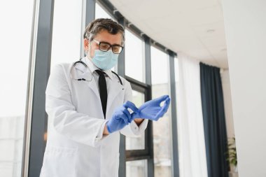 Koruyucu maskeli erkek doktor klinikte eldiven takıyor.