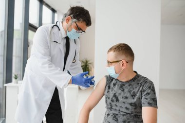 İlaç, aşı ve sağlık konsepti - virüs hastalığından korunmak için yüz koruyucu maske takan doktor ve şırınga ile erkek hastaya aşı enjekte ediliyor