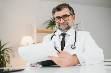 İş yerinde oturan, gülümseyerek gülümseyen yetişkin, profesyonel sağlık terapistinin portresi. Kendine güveni tam yaşlı bir erkek doktor beyaz önlüklü steteskop takıp kameraya bakıyor..