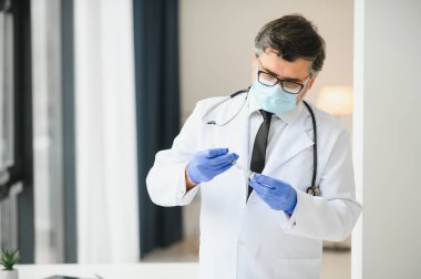 Koruyucu maske takan orta yaşlı erkek doktor elinde aşıyla bir şırınga tutuyor.