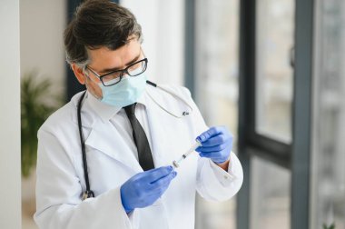 Koruyucu maske takan orta yaşlı erkek doktor elinde aşıyla bir şırınga tutuyor.