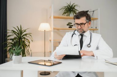 İş yerinde oturan, gülümseyerek gülümseyen yetişkin, profesyonel sağlık terapistinin portresi. Kendine güveni tam yaşlı bir erkek doktor beyaz önlüklü steteskop takıp kameraya bakıyor..