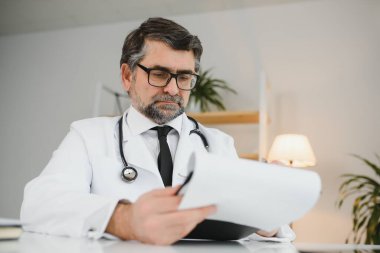 Ciddi profesyonel kıdemli yaşlı doktor iş yerindeki tıbbi belgeleri kontrol ediyor. Hasta teşhisini ya da hastane raporunu inceleyen, tıbbi form okuyan konsantre yaşlı bir doktor..