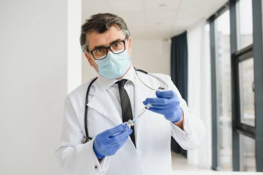 Koruyucu maske takan orta yaşlı erkek doktor elinde aşıyla bir şırınga tutuyor.