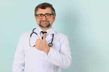 Kıdemli doktor steteskop ve mavi arka plan üzerine tıbbi ceket giyiyor. Neşeli bir ifade.