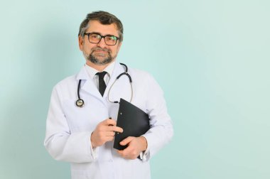 Kıdemli doktor steteskop ve mavi arka plan üzerine tıbbi ceket giyiyor. Neşeli bir ifade.