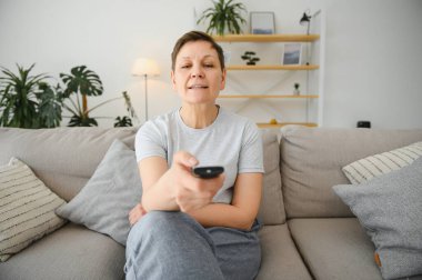 Orta yaşlı bir kadın kanepede oturuyor ve uzaktan kumandayla televizyonu açıyor.