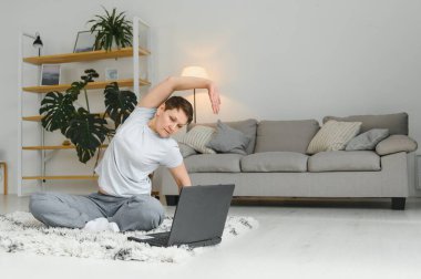 Evde internet üzerinden spor yapmak. Spor kıyafetleri içinde orta yaşlı bir kadın spor minderinde yoga yapıyor. Laptopuyla oturma odasında. İçi cihazlarla dolu.