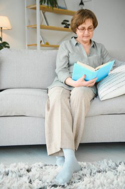 Dinlendirici okuma. Düşünceli portresi yaşlı kadın rahat ev favori edebiyat okuma. Rahat kanepe yastık üzerinde yalan söylüyor. Bacaklar yumuşak battaniye ile örtülü