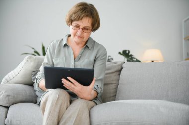 Yaşlı kadın evdeki oturma odasında dijital tablet kullanıyor..