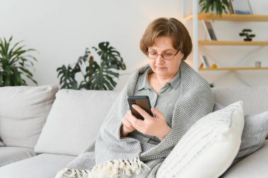 Yetişkin bir kadın akıllı telefon kullanıyor. Evdeki koltukta oturuyor. Gözlüklü, cep telefonuyla internette sörf yapan neşeli bir büyükanne. Teknoloji konsepti.