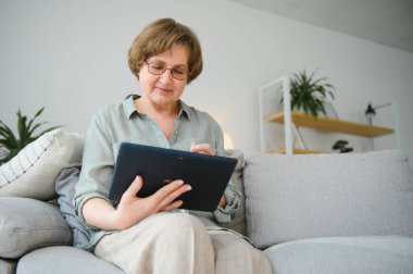 Yaşlı kadın evdeki oturma odasında dijital tablet kullanıyor..