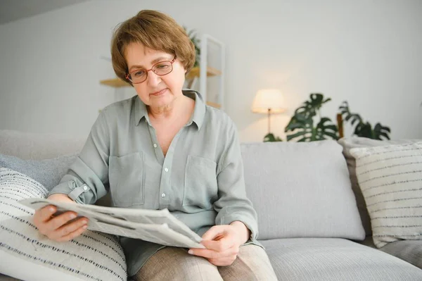 高级女士读她的报纸在家里在沙发上放松和瞥查看器顶部 — 图库照片