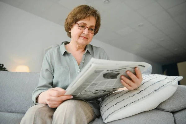 高级女士读她的报纸在家里在沙发上放松和瞥查看器顶部 — 图库照片