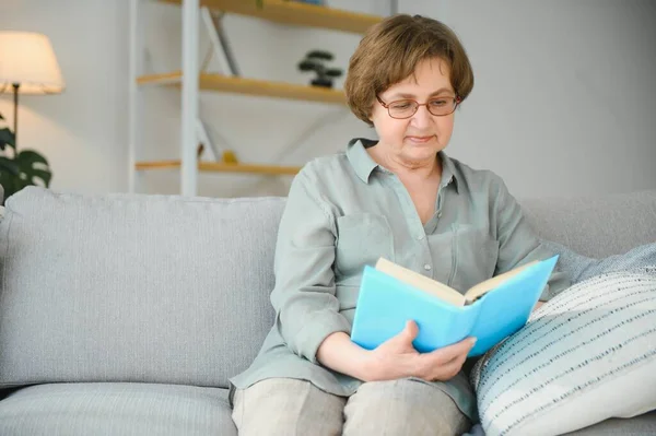宁静的阅读 在温馨的家中读喜爱文学的老年妇女画像 她躺在舒适的沙发上的枕头上 腿上覆盖着柔软的毯子 — 图库照片