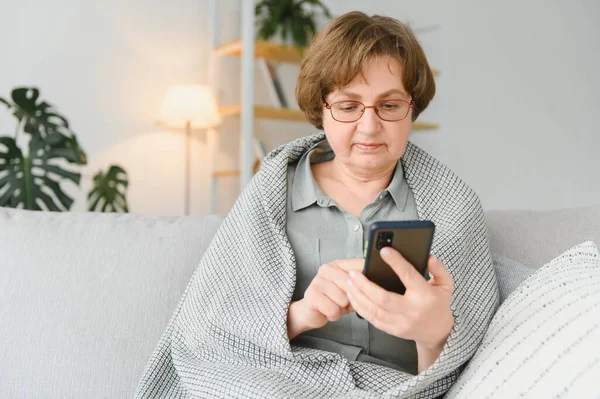坐在家里沙发上 穿着休闲装的漂亮老妇人用她的智能手机 — 图库照片