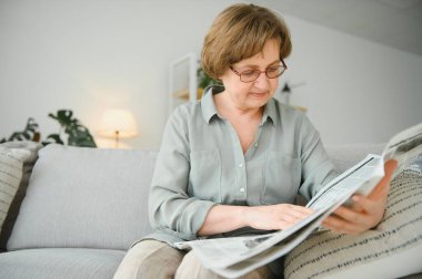 üst düzey kadın evde onun gazete okuyan bir kanepede rahatlatıcı ve görüntüleyicinin üst üzerinden bakıyor.