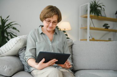 Yaşlı kadın evdeki oturma odasında dijital tablet kullanıyor..