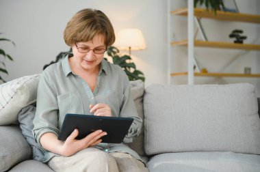 Emeklilik. Gülümseyen yaşlı kadın evdeki koltukta dijital tabletle dinleniyor, internete bakıyor ya da film izliyor..