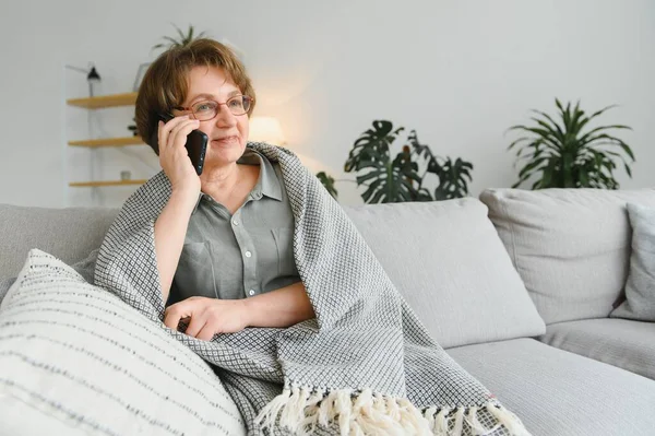 坐在家里沙发上 穿着休闲装的漂亮老妇人用她的智能手机 — 图库照片