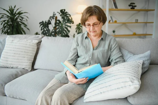 Dinlendirici okuma. Düşünceli portresi yaşlı kadın rahat ev favori edebiyat okuma. Rahat kanepe yastık üzerinde yalan söylüyor. Bacaklar yumuşak battaniye ile örtülü