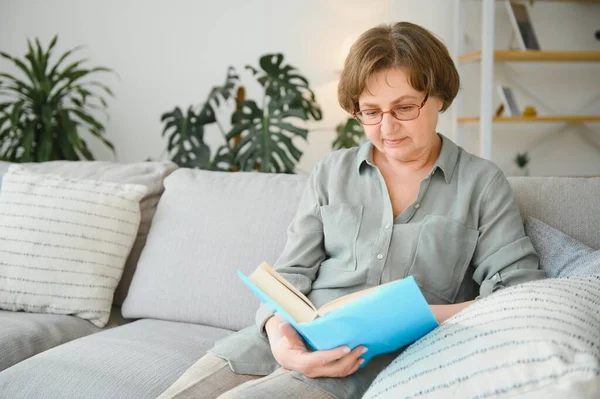 Dinlendirici okuma. Düşünceli portresi yaşlı kadın rahat ev favori edebiyat okuma. Rahat kanepe yastık üzerinde yalan söylüyor. Bacaklar yumuşak battaniye ile örtülü